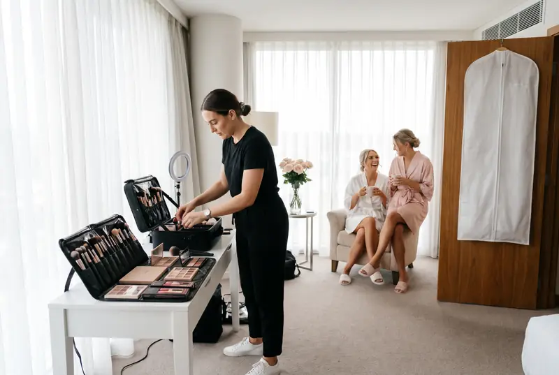 A makeup artist sets up her kit at a table in a bridal suite while the bride laughs by the window