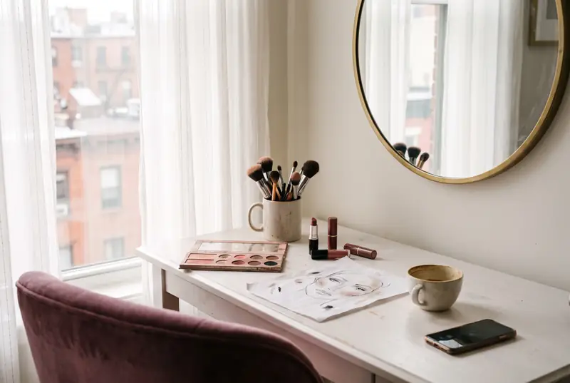 A makeup artist's vanity by a window: brushes in a cup, an open palette, a face chart and a coffee